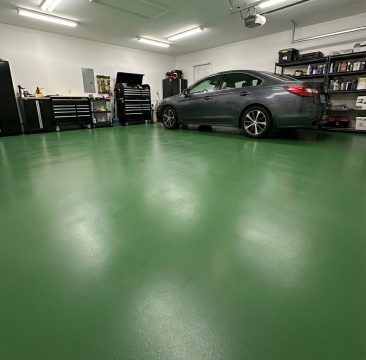 Jasno oświetlony garaż z lśniącą, epoksydową podłogą w kolorze RAL 6001 (zielony mech) zajmuje centralną część obrazu.