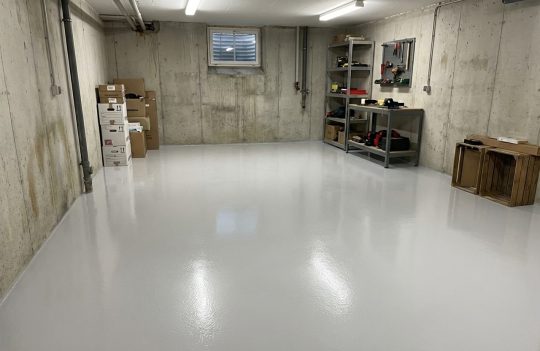 Gładka, lśniąca posadzka w kolorze RAL 7038 wypełnia całe przestronne pomieszczenie piwniczne o surowych, betonowych ścianach.