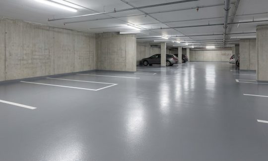 Przestronny parking podziemny z lśniącą posadzką w kolorze szarym, RAL 7046, oraz białymi liniami wyznaczającymi miejsca postojowe. Surowe betonowe ściany i filary dopełniają industrialnego wyglądu, a w tle widać kilka zaparkowanych samochodów.
